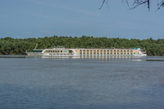View of a large river cruise ship sailing on the Danube river with a lush green forest background under a clear blue sky in Budapest Hungary.