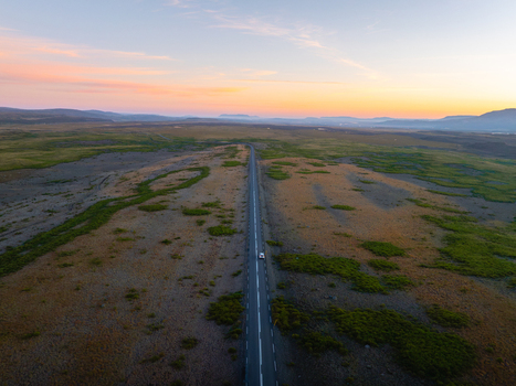 Aerial view of a single road cutting through a vast landscape the sky painted with hues of orange and blue Reykholt Iceland.