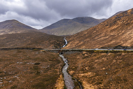 Aerial view of Eas a Bhradain waterfall cascading down rugged mountain slopes into a stream crossing under a road in the moorland landscape Isle of Skye Scotland United Kingdom. Aerial view of Eas a Bhradain waterfall cascading down rugged mountain slopes into a stream crossing under a road in the moorland landscape Isle of Skye Scotland United Kingdom.