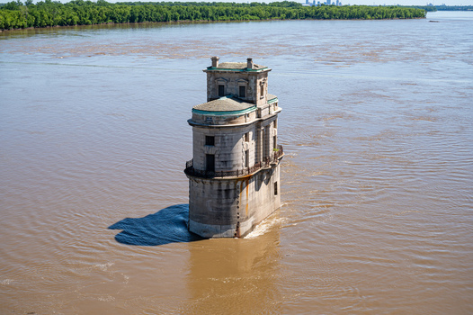 View of the historic water intake tower rising from the Mississippi River with calm brown water and lush green trees along the distant riverbank in St. Louis Missouri United States. View of the historic water intake tower rising from the Mississippi River with calm brown water and lush green trees along the distant riverbank in St. Louis Missouri United States.