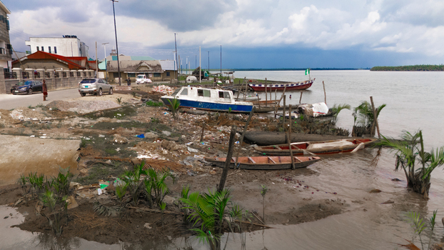 Aerial view of wooden boats and a small vessel moored along a muddy riverbank with scattered debris palm trees and buildings in Rivers Nigeria.