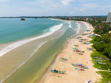 Aerial view of traditional fishing boats lined up on the sandy beach with turquoise ocean waves and lush palm trees along the coast in Weligama Southern Province Sri Lanka.