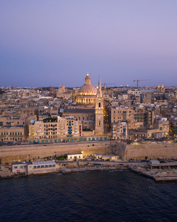 Aerial view of St. Paul s Pro Cathedral with its iconic spire and dome rising above historic stone buildings at dusk overlooking the Mediterranean Sea in Valletta Port Region Malta. Aerial view of St. Paul s Pro Cathedral with its iconic spire and dome rising above historic stone buildings at dusk overlooking the Mediterranean Sea in Valletta Port Region Malta.