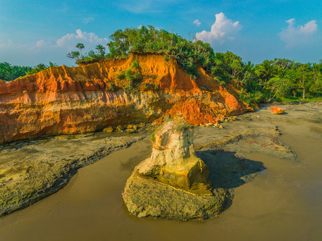 Aerial view of a dramatic coastal cliff with orange hued rock formations lush green vegetation on top and dark sand beach in Bali Indonesia. Aerial view of a dramatic coastal cliff with orange hued rock formations lush green vegetation on top and dark sand beach in Bali Indonesia.