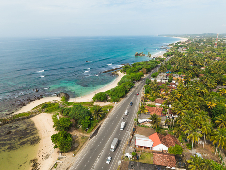 Aerial view of a coastal road with vehicles running along the sandy beach and turquoise ocean waves under a clear sky in Koggala Southern Province Sri Lanka. Aerial view of a coastal road with vehicles running along the sandy beach and turquoise ocean waves under a clear sky in Koggala Southern Province Sri Lanka.