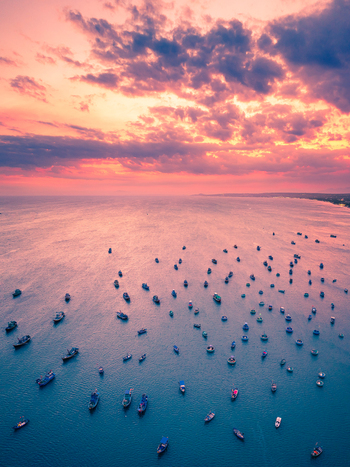 Aerial view of numerous small fishing boats scattered across the calm blue sea under a vibrant pink and orange sunset sky in Vietnam. Aerial view of numerous small fishing boats scattered across the calm blue sea under a vibrant pink and orange sunset sky in Vietnam.