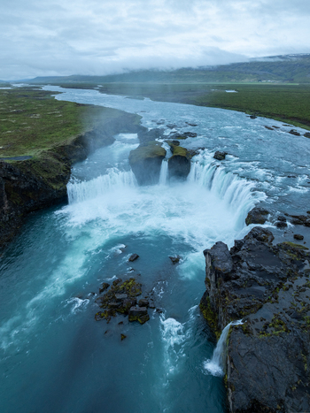 Aerial view of Godafoss Waterfall flowing over dark volcanic rocks into a turquoise river surrounded by lush green mossy landscape under a cloudy sky in Thingeyjarsveit Iceland. Aerial view of Godafoss Waterfall flowing over dark volcanic rocks into a turquoise river surrounded by lush green mossy landscape under a cloudy sky in Thingeyjarsveit Iceland.