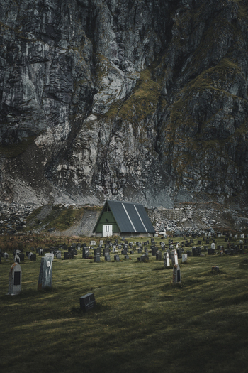 View of Flakstad Church with its green roof and white doors surrounded by a cemetery with weathered gravestones set against a towering rugged mountain cliff in Ramberg Nordland Norway. View of Flakstad Church with its green roof and white doors surrounded by a cemetery with weathered gravestones set against a towering rugged mountain cliff in Ramberg Nordland Norway.