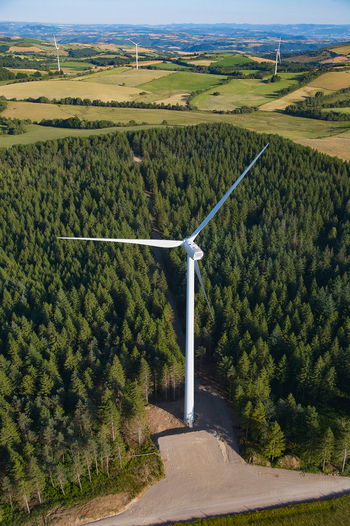 Aerial view of a towering wind turbine standing amidst a dense forest juxtaposed against rolling green hills under a clear sky Salles Curan Occitanie France. Aerial view of a towering wind turbine standing amidst a dense forest juxtaposed against rolling green hills under a clear sky Salles Curan Occitanie France.