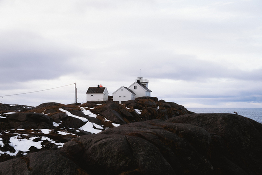 View of stark white buildings perch atop rugged dark rocks dusted with snow against the moody overcast sky in Henningsv&aelig;r Nordland Norway.