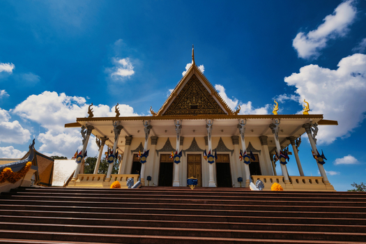 View of the Royal Palace of Cambodia featuring ornate golden roof architecture white pillars and grand stairs under a bright blue sky in Phnom Penh Phnom Penh Cambodia. View of the Royal Palace of Cambodia featuring ornate golden roof architecture white pillars and grand stairs under a bright blue sky in Phnom Penh Phnom Penh Cambodia.