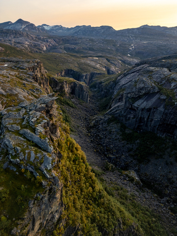 Aerial view of the deep Hellmojuvet canyon with steep rocky cliffs sparse vegetation and distant mountain peaks under a warm golden hour sky in Hamaroy Nordland Norway.