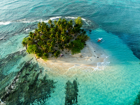 Aerial view of Guyam Island featuring lush palm trees a white sand beach and clear turquoise water with a small boat anchored nearby in General Luna Caraga Philippines. Aerial view of Guyam Island featuring lush palm trees a white sand beach and clear turquoise water with a small boat anchored nearby in General Luna Caraga Philippines.