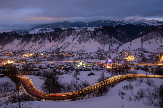 View of the village of Moieciu de Sus at night with snow covered mountains glowing house lights and light trails from vehicles on a winding road in Moieciu de Sus Brasov Romania. View of the village of Moieciu de Sus at night with snow covered mountains glowing house lights and light trails from vehicles on a winding road in Moieciu de Sus Brasov Romania.