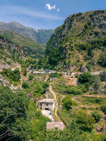Aerial view of a traditional stone building nestled in a lush green valley surrounded by rugged mountains and a winding dirt path under a clear blue sky in Dhermi Vlore County Albania.