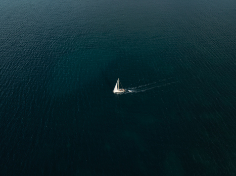 Aerial view of a lone sailboat with white sails gliding across the deep blue Mediterranean Sea near Syracuse Sicily Italy. Aerial view of a lone sailboat with white sails gliding across the deep blue Mediterranean Sea near Syracuse Sicily Italy.