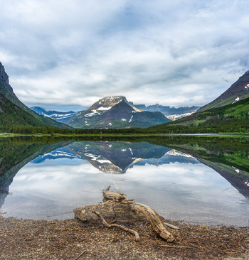 View of the snow capped peaks of Glacier National Park reflecting in the still waters of a mountain lake with driftwood on the shore in Montana United States. View of the snow capped peaks of Glacier National Park reflecting in the still waters of a mountain lake with driftwood on the shore in Montana United States.