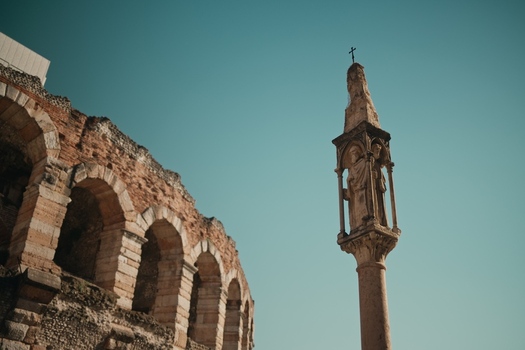 View of the ancient Roman Arena and a historic stone column with a statue under a clear blue sky in Verona Veneto Italy.