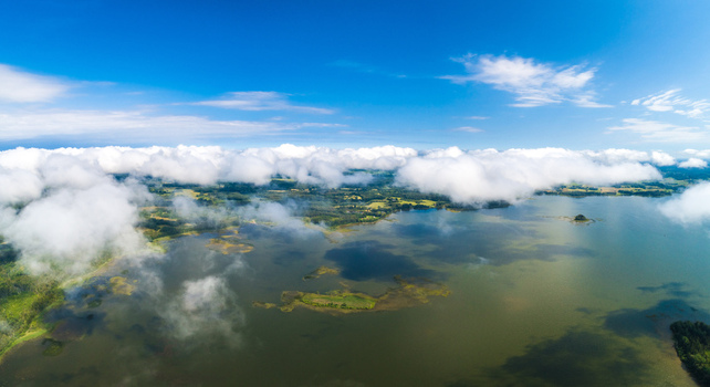 Aerial view of a serene lake surrounded by lush green forests and fields under a bright blue sky with scattered white clouds in Latvia. Aerial view of a serene lake surrounded by lush green forests and fields under a bright blue sky with scattered white clouds in Latvia.