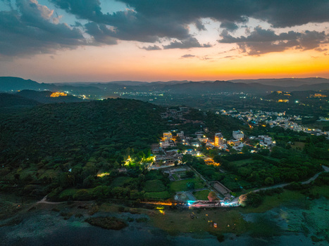 Aerial view of the landscape surrounding the city at sunset featuring rolling hills illuminated buildings and the calm waters of a lake in Udaipur Rajasthan India.