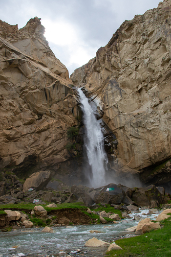 View of Khamosh Waterfall cascading down steep rocky cliffs into a stream surrounded by rugged mountain terrain and green grass in Skardu Gilgit Pakistan.