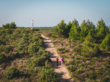 Aerial view of a winding path leading up a grassy hill towards a white cross framed by lush greenery under a clear sky Istria County Croatia.