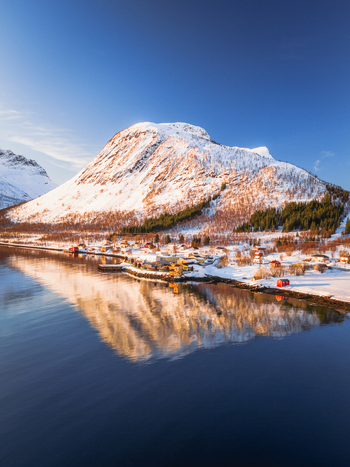 Aerial view of snow capped mountains mirroring in the tranquil waters contrasting with the small village nestled along the shore Indregård Troms Norway. Aerial view of snow capped mountains mirroring in the tranquil waters contrasting with the small village nestled along the shore Indregård Troms Norway.