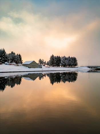 Aerial view of a snowy Arctic landscape at sunset with a small building and snow covered trees reflected in the calm water in Tromso Troms Norway. Aerial view of a snowy Arctic landscape at sunset with a small building and snow covered trees reflected in the calm water in Tromso Troms Norway.