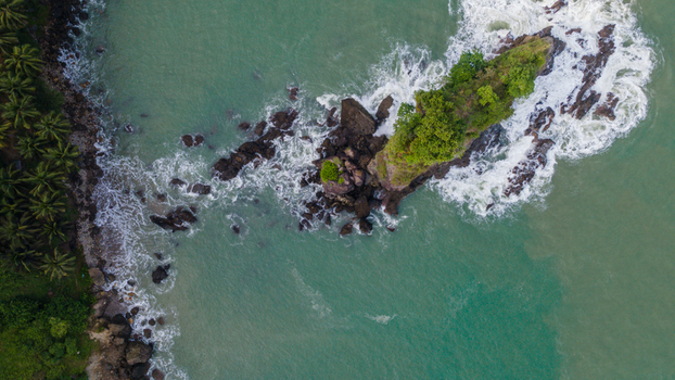 Aerial view of Pantai Karang Agung featuring a rocky outcrop with green trees surrounded by turquoise ocean waves and a palm fringed coast in Ayah Central Java Indonesia. Aerial view of Pantai Karang Agung featuring a rocky outcrop with green trees surrounded by turquoise ocean waves and a palm fringed coast in Ayah Central Java Indonesia.