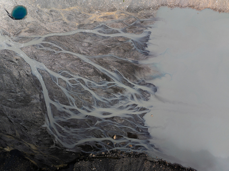 Aerial view of a braided glacial river with sediment rich water flowing through dark rocky terrain and a small blue glacial pool in Nepal.