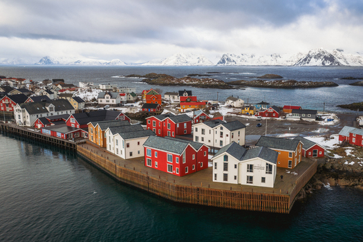 Aerial view of vibrant red and white houses cluster tightly along the dark pier against a backdrop of snow dusted mountains and icy waters Henningsv&aelig;r Nordland Norway.