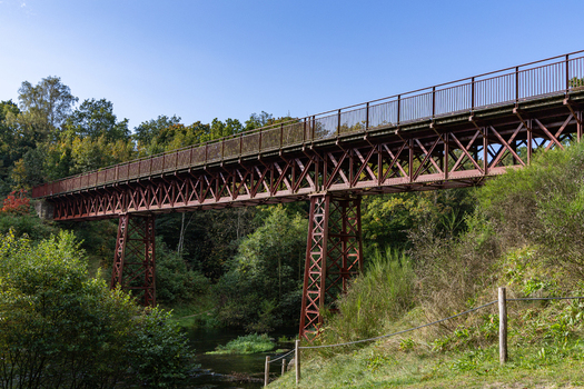 View of The Uncovered Bridge a historic red steel truss structure spanning a river surrounded by lush green forest in Braedstrup Jutland Denmark. View of The Uncovered Bridge a historic red steel truss structure spanning a river surrounded by lush green forest in Braedstrup Jutland Denmark.