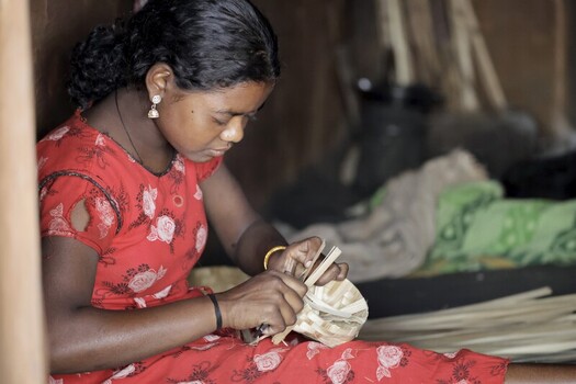 Odisha India 26 March 2026 View of a young woman dressed in a vibrant red floral dress meticulously weaving a basket with focused concentration. Odisha India 26 March 2026 View of a young woman dressed in a vibrant red floral dress meticulously weaving a basket with focused concentration.