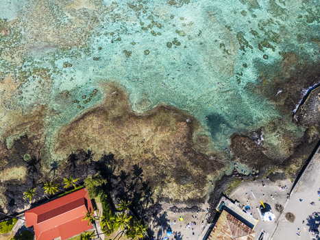 Aerial view of Kahalu u Beach Park featuring turquoise waters coral reefs and people relaxing on the sandy shore with palm trees and a red roofed building Kailua Kona Hawaii United States. Aerial view of Kahalu u Beach Park featuring turquoise waters coral reefs and people relaxing on the sandy shore with palm trees and a red roofed building Kailua Kona Hawaii United States.