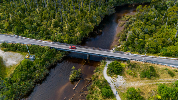 Aerial view of a lone red car crossing a bridge over a dark river surrounded by a dense green forest Ship Creek Haast West Coast Region New Zealand. Aerial view of a lone red car crossing a bridge over a dark river surrounded by a dense green forest Ship Creek Haast West Coast Region New Zealand.