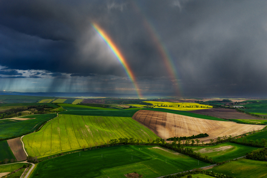 Aerial view of a double rainbow arching over vibrant green and brown agricultural fields under a dramatic stormy sky with rain showers in Nasedlovice South Moravian Region Czechia.