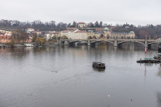 View of the Vltava River with a boat cruising past the Manes Bridge and the Straka Academy building under an overcast sky in Prague Prague Czechia. View of the Vltava River with a boat cruising past the Manes Bridge and the Straka Academy building under an overcast sky in Prague Prague Czechia.