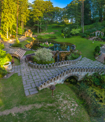 Aerial view of a lush green park featuring a tranquil pond with a small island a stone bridge with decorative arches and manicured garden paths in Latvia. Aerial view of a lush green park featuring a tranquil pond with a small island a stone bridge with decorative arches and manicured garden paths in Latvia.