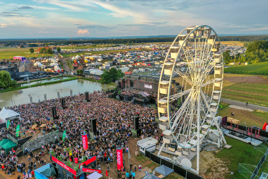 Herzogenaurach Germany 24 July 2025 Aerial view of Open Beatz Festival featuring a large crowd a white Ferris wheel and a main stage next to a pond under a bright sky.