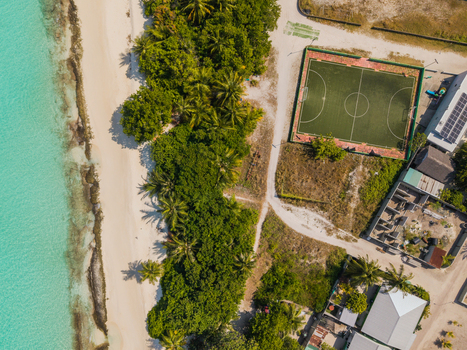 Aerial view of turquoise waters kissing the white sandy beach and lush green vegetation contrasting with the football field Vashafaru Island Thiladhummathi Atoll Maldives.