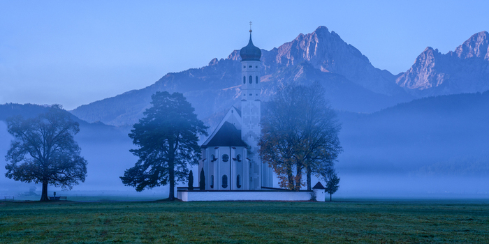 Aerial view of St. Coloman Church surrounded by a misty field with mountains in the background in Germany. Aerial view of St. Coloman Church surrounded by a misty field with mountains in the background in Germany.