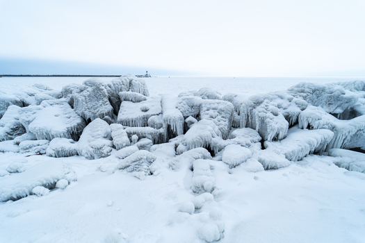 View of the frozen breakwater rocks covered in thick ice and icicles with the distant Molenfeuer lighthouse visible against a cold sky in Sassnitz Mecklenburg Vorpommern Germany.