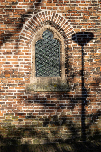 View of the historic arched window of the Sint Nicolaaskapel with a metal grate and a street lamp shadow on the brick wall in Nijmegen Gelderland Netherlands. View of the historic arched window of the Sint Nicolaaskapel with a metal grate and a street lamp shadow on the brick wall in Nijmegen Gelderland Netherlands.