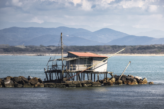 View of a traditional wooden fishing hut on a rocky pier with the turquoise sea and distant mountains in Marina di Pisa Tuscany Italy.
