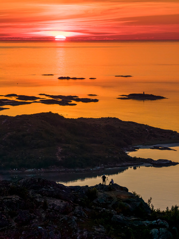 Aerial view of Sommaroy featuring rocky coastal terrain small islands and calm water under a vibrant sunset sky with two photographers on a cliff in Sommaroy Troms Norway. Aerial view of Sommaroy featuring rocky coastal terrain small islands and calm water under a vibrant sunset sky with two photographers on a cliff in Sommaroy Troms Norway.