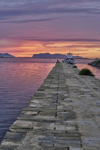 View of the historic Saline dello Stagnone salt pans at sunset with a stone pier leading towards a boat on the calm water in Marsala Sicilia Italy.