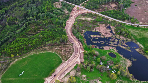 Aerial view of a winding dirt access road cutting through rural forests and green fields near a dark pond in Latvija. Aerial view of a winding dirt access road cutting through rural forests and green fields near a dark pond in Latvija.