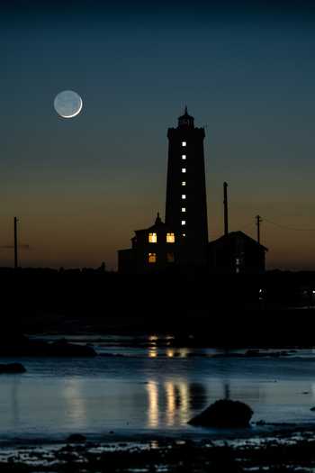 View of Grotta Lighthouse silhouetted against a twilight sky with a crescent moon and glowing windows in Seltjarnarnes Seltjarnarnesbaer Iceland. View of Grotta Lighthouse silhouetted against a twilight sky with a crescent moon and glowing windows in Seltjarnarnes Seltjarnarnesbaer Iceland.