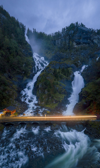 Aerial view of Latefossen twin waterfalls with car light trails on the stone bridge and lush green mountains at dusk Skare Vestland Norway.