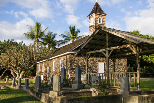 View of the historic Keawala i Congregational Church with its stone walls and cemetery in the foreground surrounded by palm trees under a blue sky in Maui County Hawaii United States.